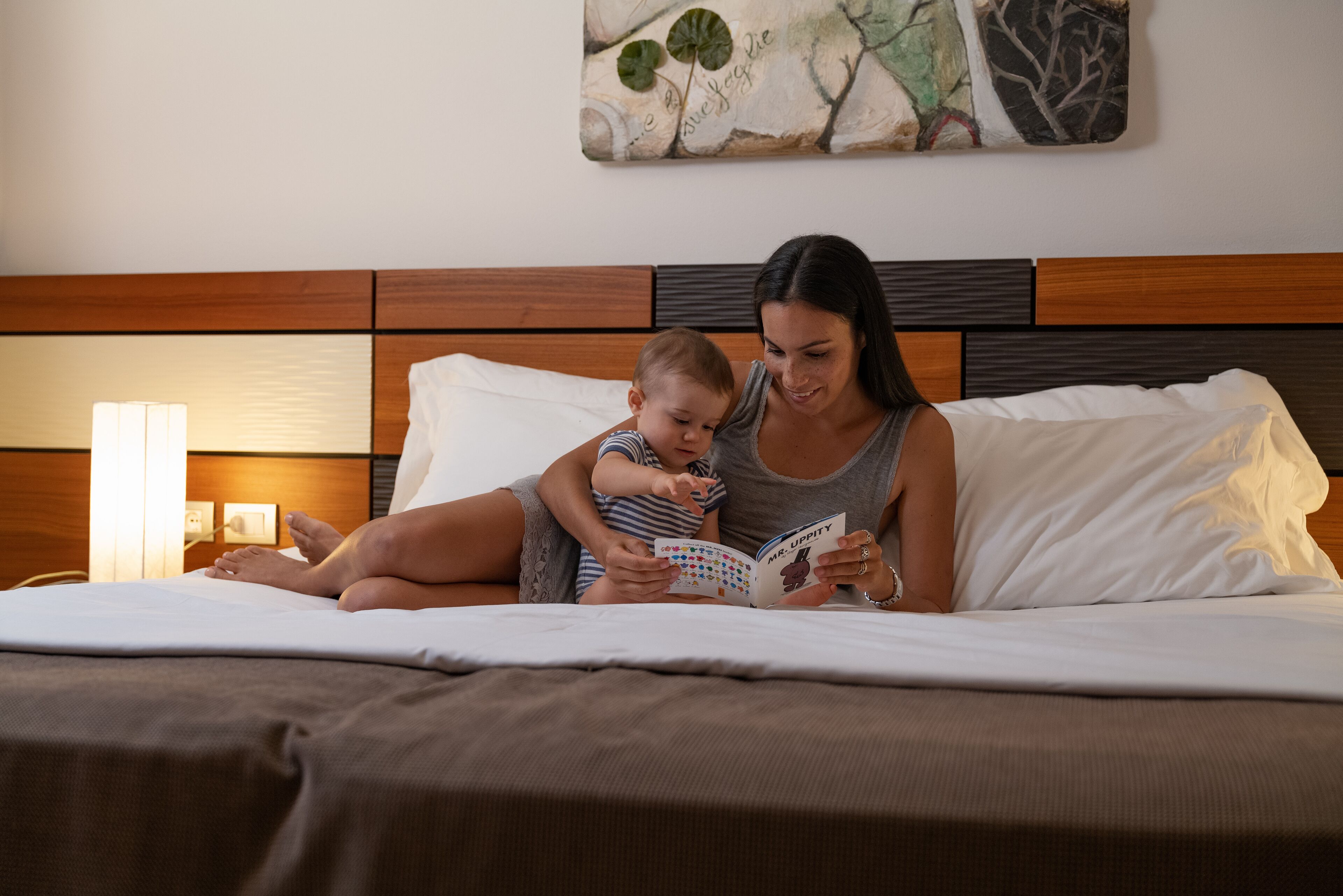 Une femme et un enfant sont assis sur un lit, devant une tête de lit en bois et des oreillers blancs, et ils lisent un livre.