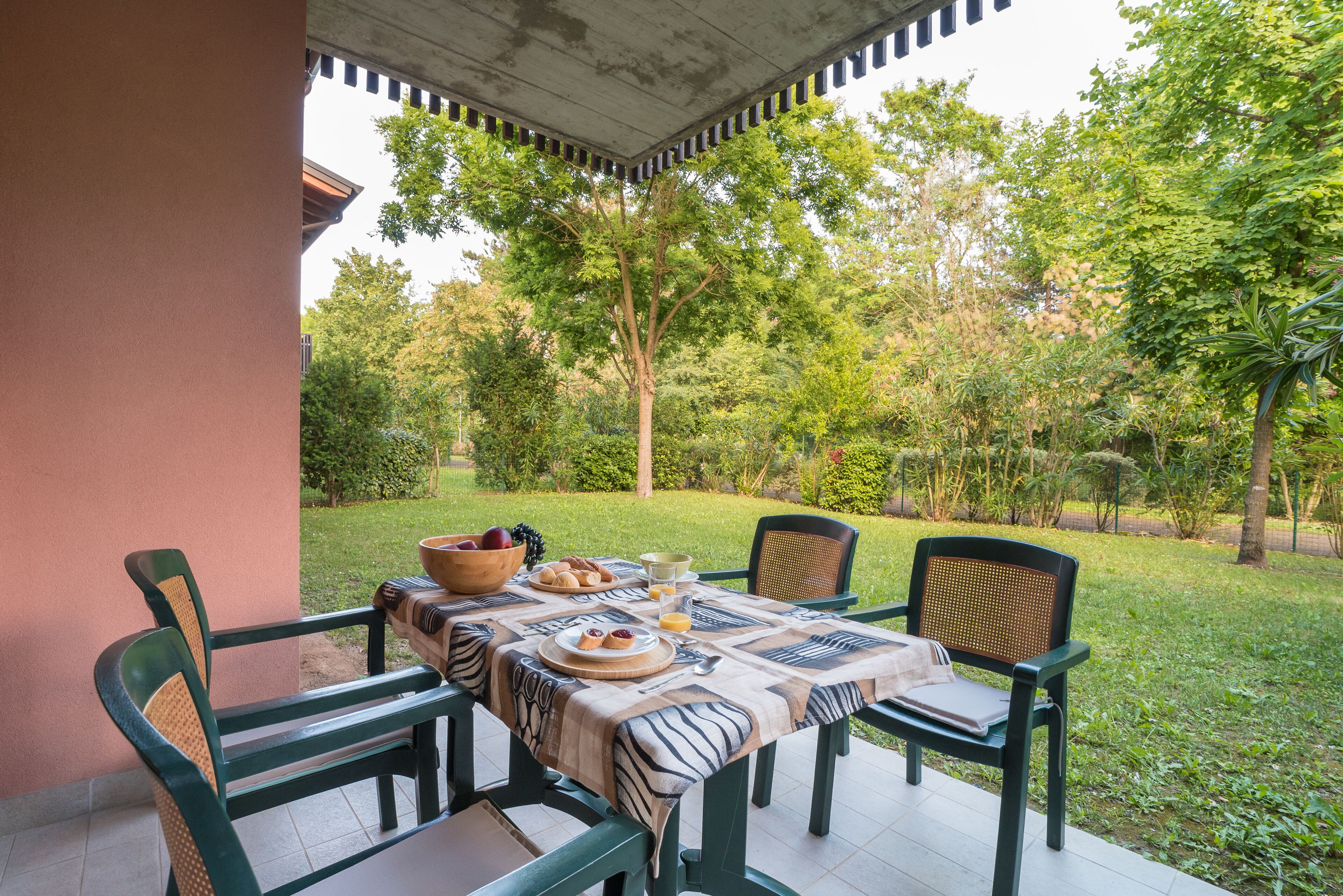 An outdoor dining area with a table set for four, surrounded by chairs, in a garden with trees and a building in the background.