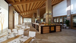 A spacious dining area with wooden tables and chairs, a central buffet station, and large windows allowing natural light. - Green Village Eco Resort (Lignano Sabbiadoro)
