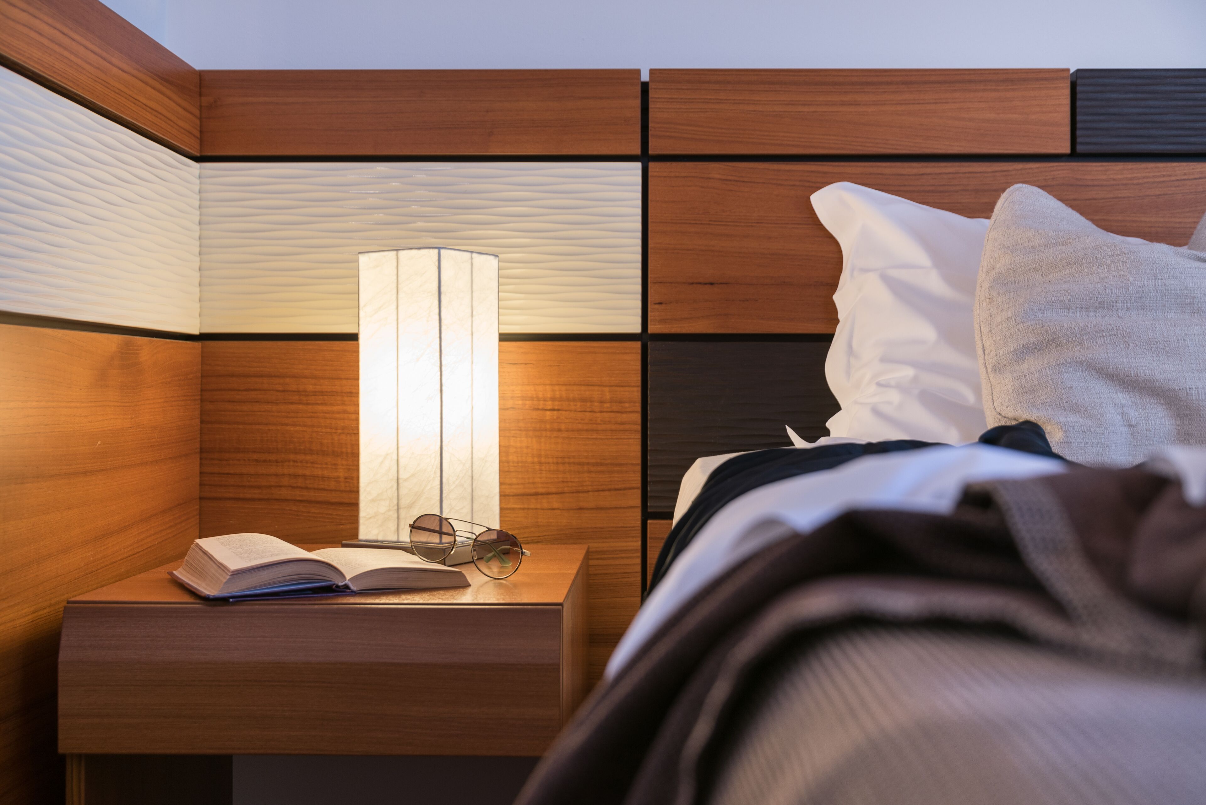 Ein ordentlich bezogenes Bett mit Kissen und einem Nachttisch mit Lampe, Buch und Brille.