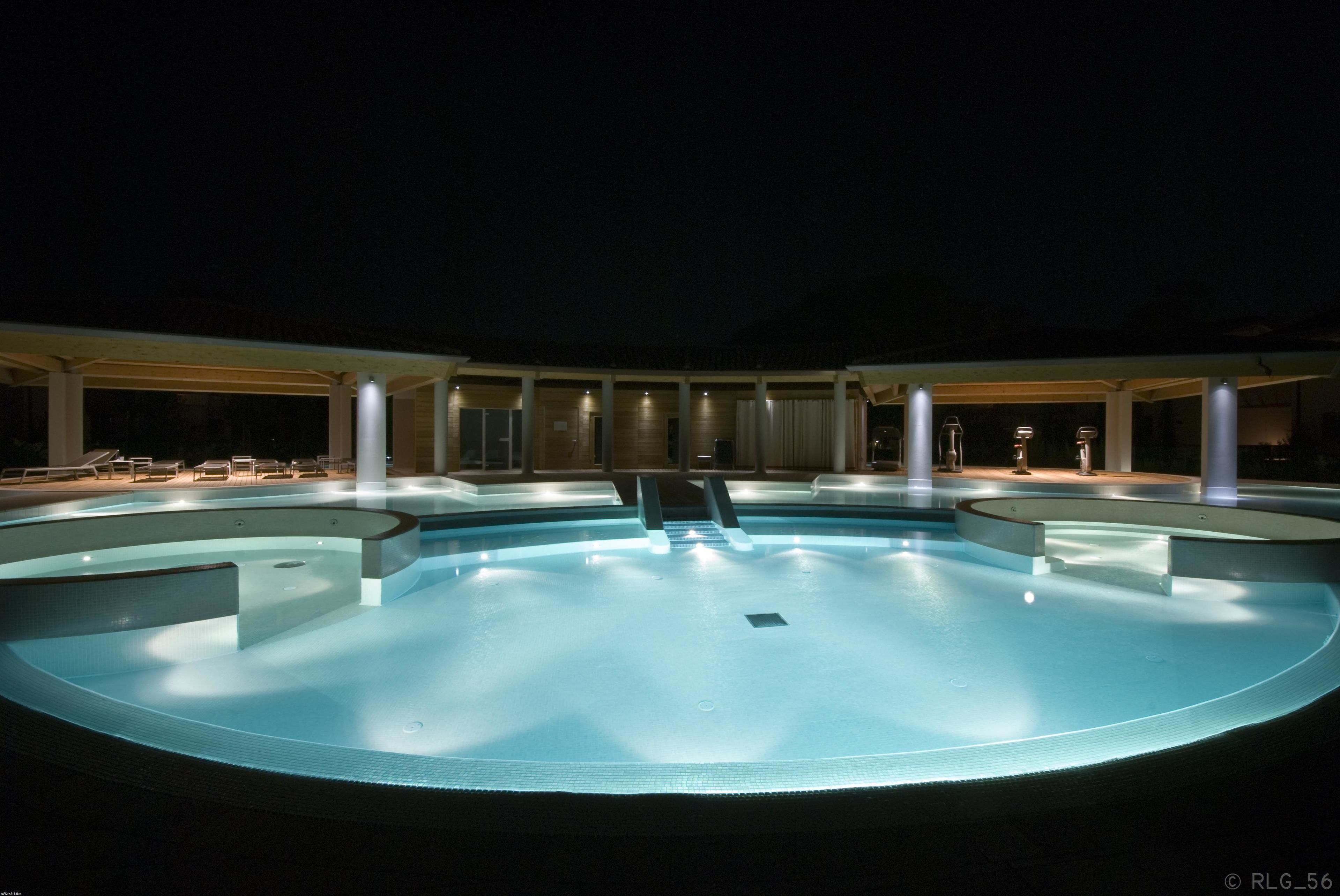 Una amplia piscina circular con un tobogán en el centro, rodeada por una zona cubierta con asientos e iluminación.