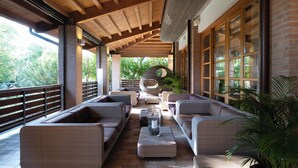 A covered outdoor seating area with wicker furniture, a glass table with a candle, and a view of greenery. - Green Village Eco Resort (Lignano Sabbiadoro)