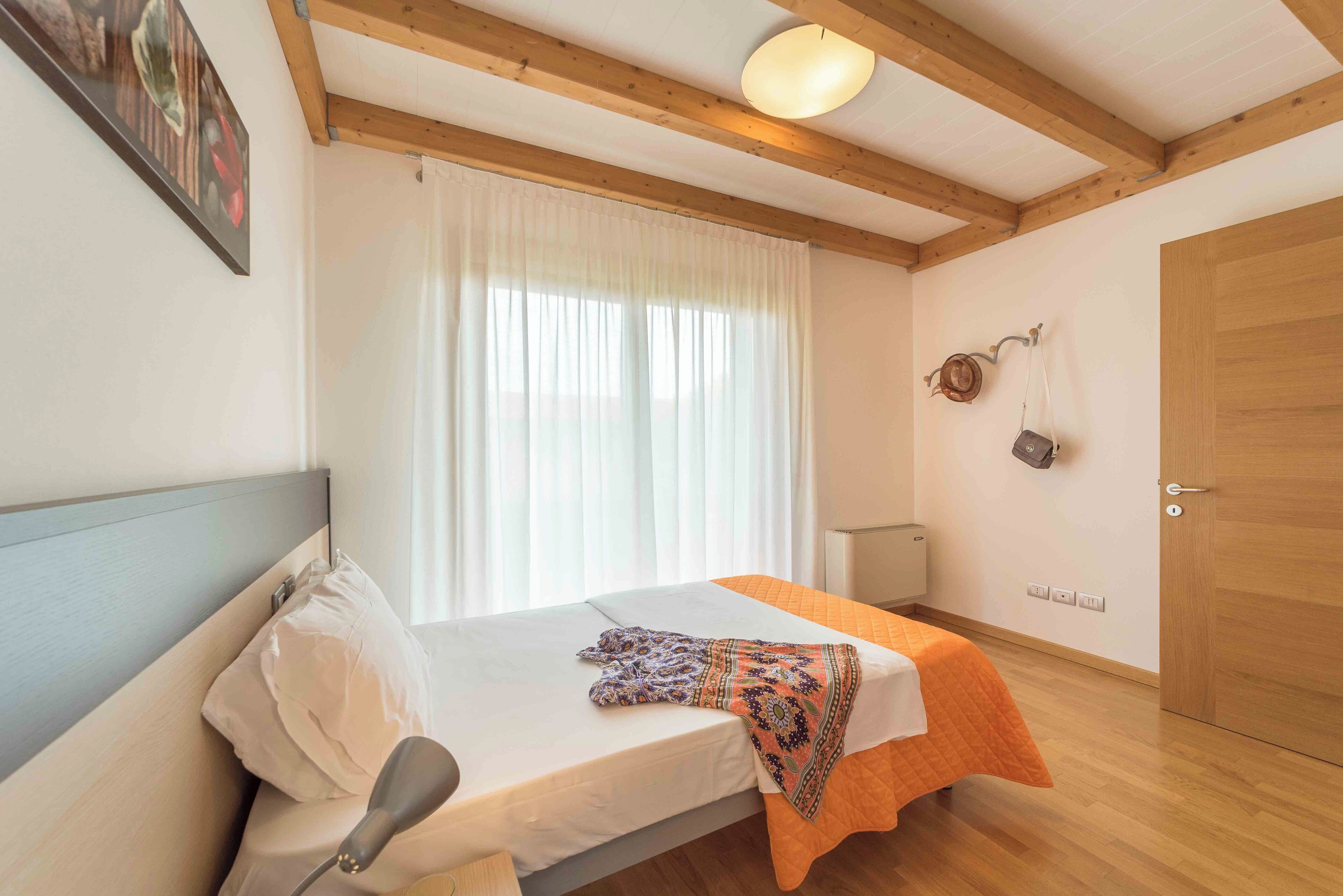 Une chambre à coucher comprenant un lit, une table de chevet, une lampe, une fenêtre avec des rideaux, une porte en bois et un porte-manteau fixé au mur.