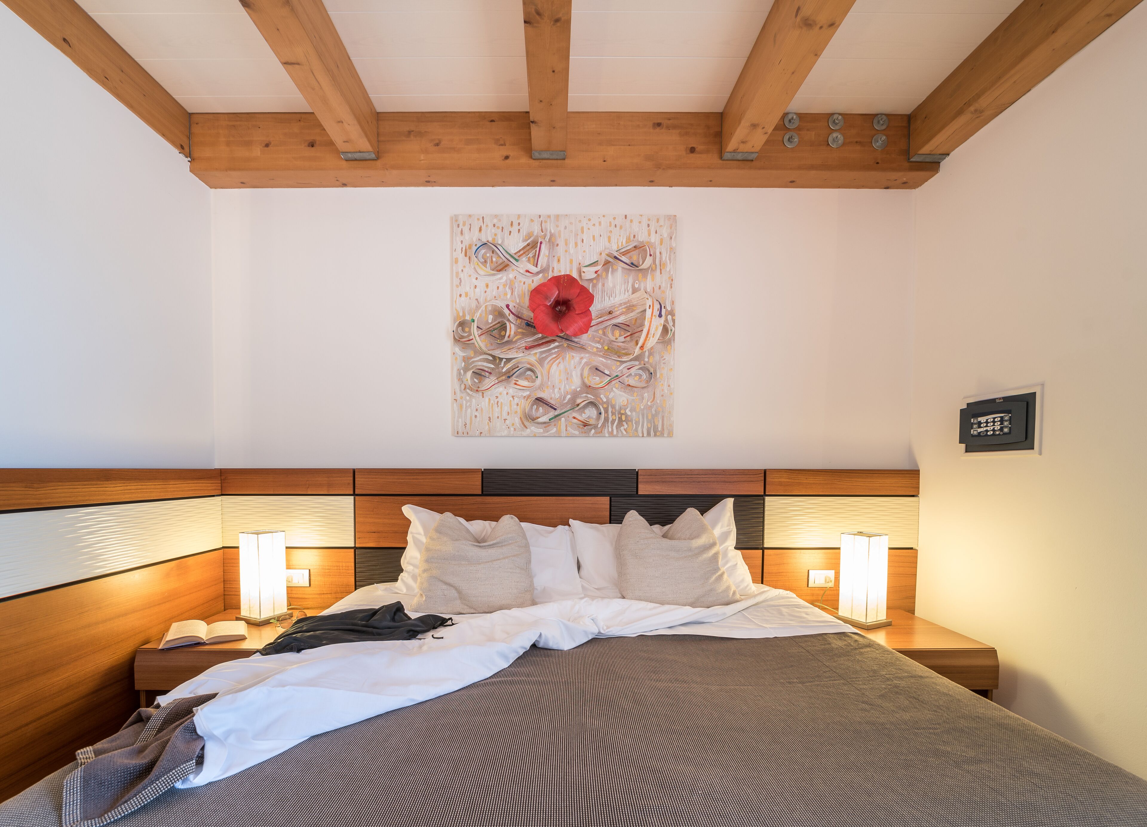 A hotel room with a wooden headboard, a large bed with white and gray bedding, two bedside lamps, and a wall-mounted artwork featuring a red flower.
