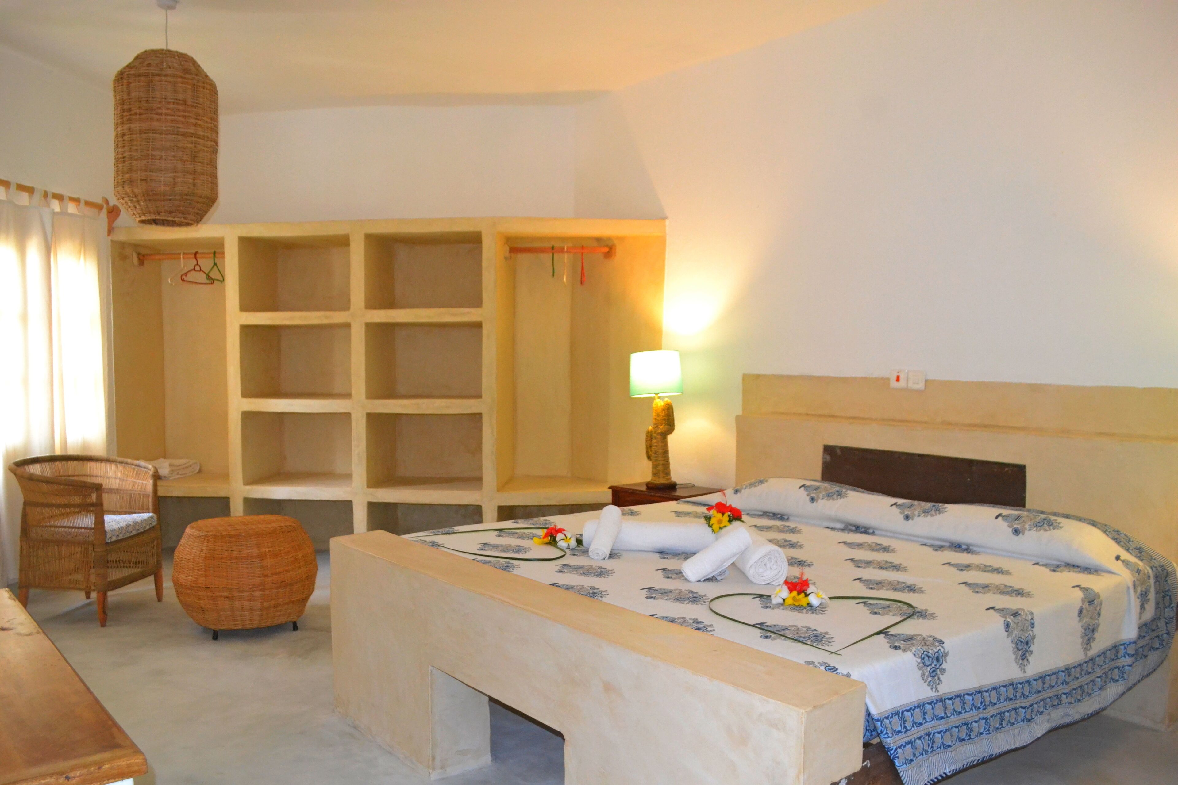 A hotel room with a bed, a wooden bench, a wicker chair, and a wardrobe with multiple shelves.