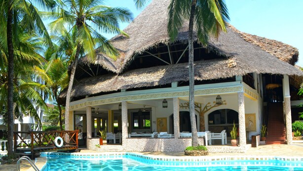 Un resort con piscina, estructura de techo de paja, palmeras y un muro de piedra.