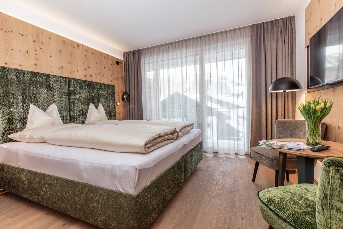 Ein modernes Hotelzimmer mit einem großen Bett, einem Holz-Kopfteil, einem Schreibtisch mit Stuhl und einem großen Fenster mit Vorhängen.