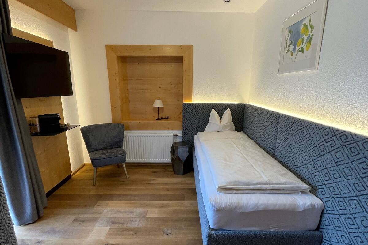 Ein kleines, gemütliches Zimmer mit einem Bett, einem Sessel, einer Heizung und einer Holzwand.
