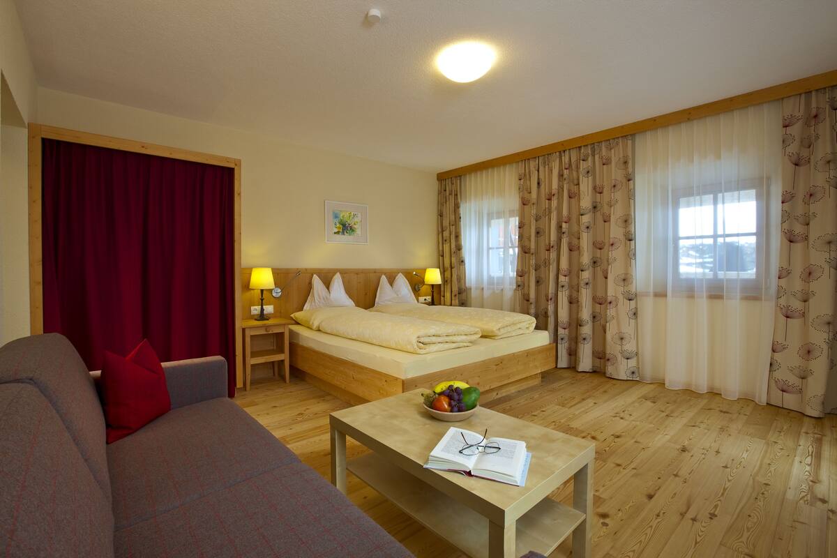 Ein Hotelzimmer mit einem Bett, einer Couch, einem Tisch mit Obstschale und Gläsern sowie Vorhängen.
