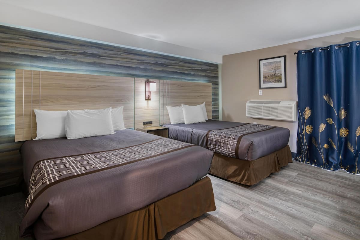 A hotel room with two beds, a large wall mural, a mounted air conditioner, and a window with a blue curtain.