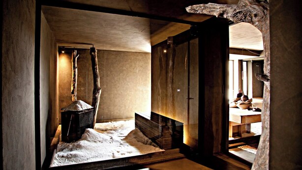 A rustic room with a wooden bench, a stone fireplace, and a view of a kitchen area.