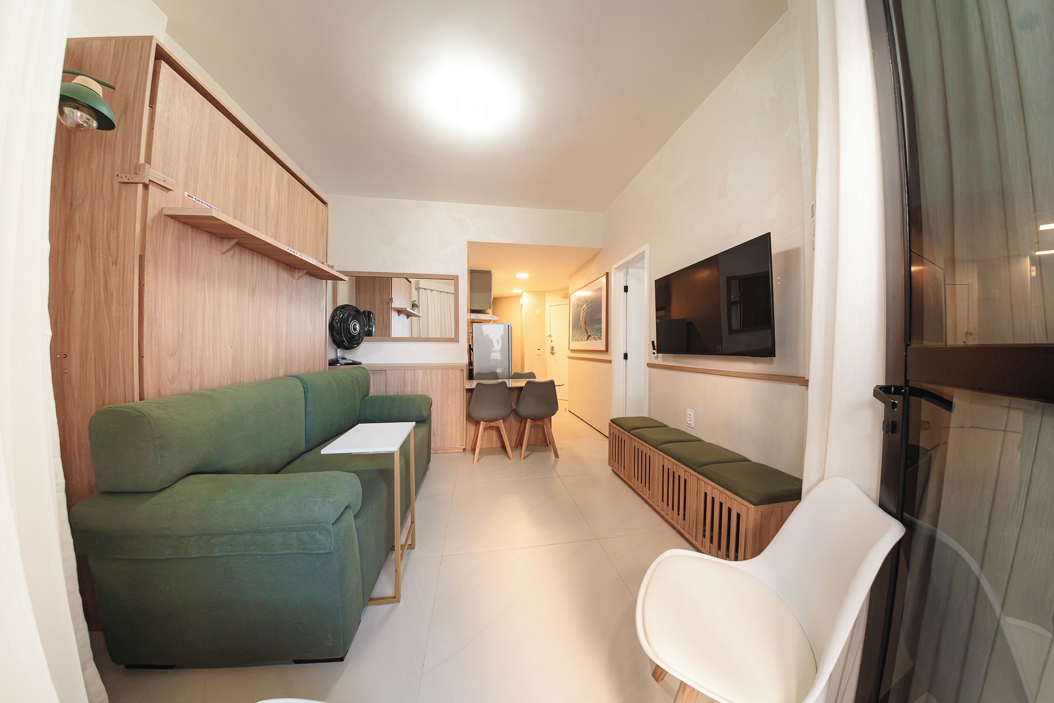 A compact living space with a green sofa, a small dining table with chairs, a TV mounted on the wall, and a kitchenette area.