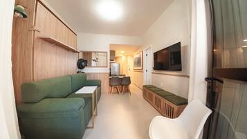 A compact living space with a green sofa, a small dining table with chairs, a TV mounted on the wall, and a kitchenette area.