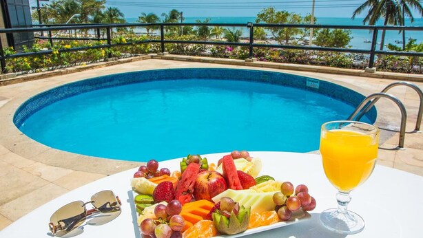 A poolside table with a fruit platter, a glass of orange juice, and sunglasses, overlooking a clear blue pool with a view of the ocean and palm trees. - Flat Golden Fortaleza (Fortaleza)