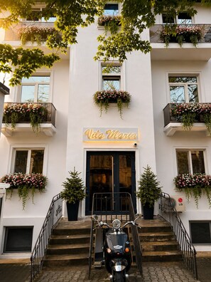 A building with a sign reading 'Vela Maria,' a scooter parked at the entrance, and flower boxes on the balconies.