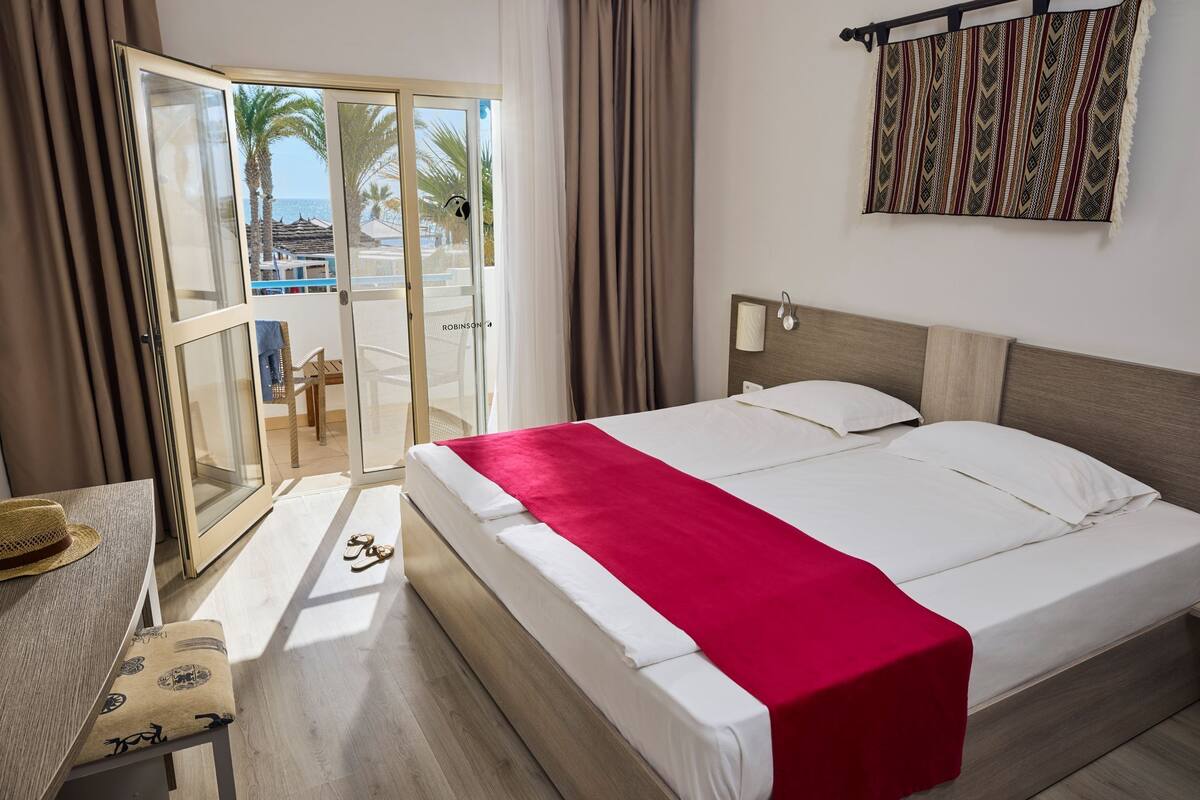 A hotel room with a bed, a desk, a chair, and a view of the beach and palm trees through the glass door.
