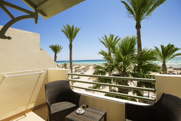 Un balcon agrémenté de meubles en osier, une petite table avec une tasse et une soucoupe, et une vue sur la plage, avec ses palmiers et ses parasols.