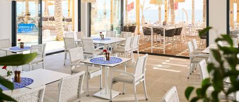 Un espace de restauration intérieur avec des meubles en osier blanc, des luminaires suspendus en fibres tressées et de grandes fenêtres offrant une vue sur la plage.