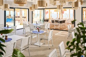 An indoor dining area with white wicker furniture, hanging woven light fixtures, and large windows offering a view of the beach.