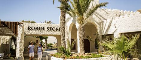 Un bâtiment avec une enseigne indiquant « ROBIN STORE » à l’entrée, entouré de palmiers et d’un jardin bien entretenu.