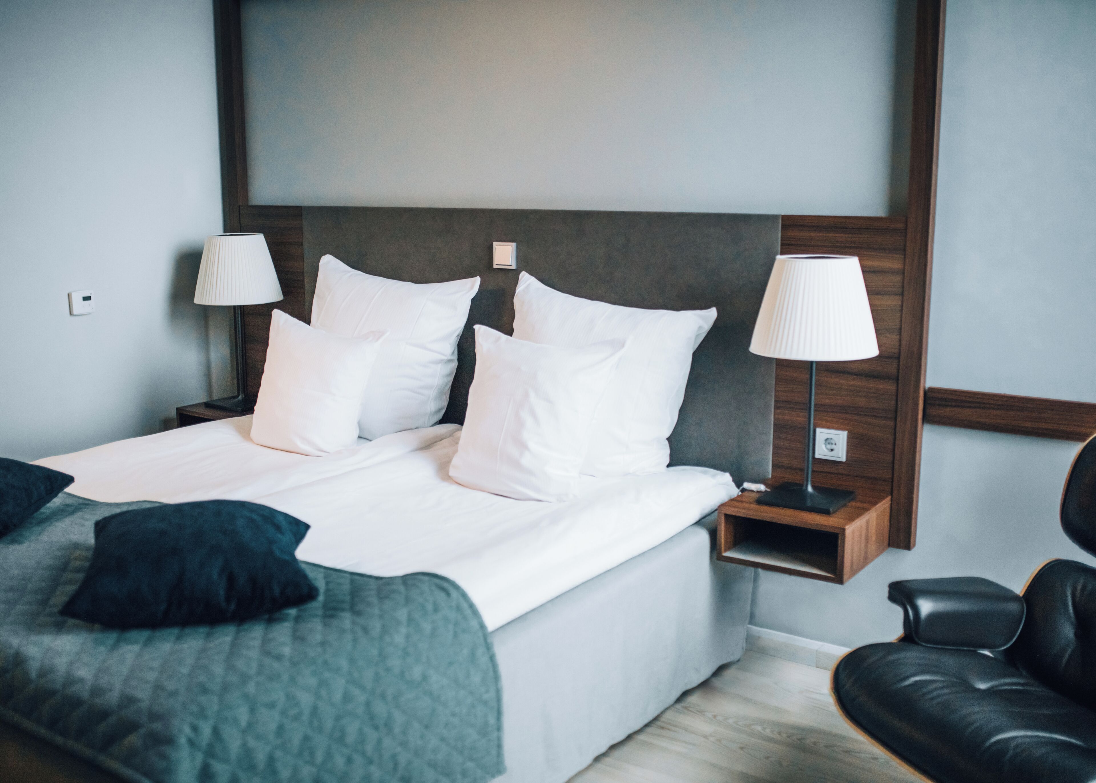 Un lit bien fait, avec du linge de lit blanc et des oreillers, une table de chevet avec une lampe, un fauteuil en cuir noir et une tête de lit en bois.