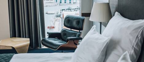 Una habitación de hotel con una cama, un sillón de cuero, una lámpara y vista al puerto.