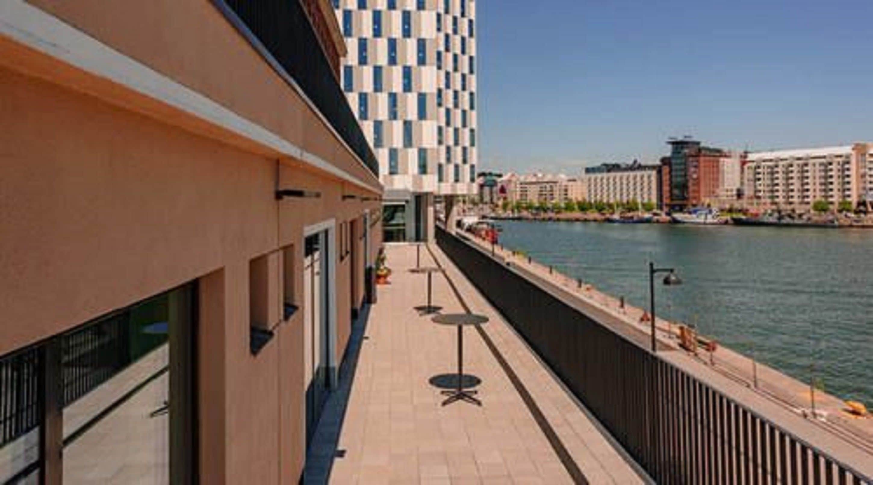 Un bâtiment moderne à la façade à motifs géométriques, une promenade au bord de la rivière avec des espaces de repos en plein air, et une vue sur le paysage urbain en arrière-plan.