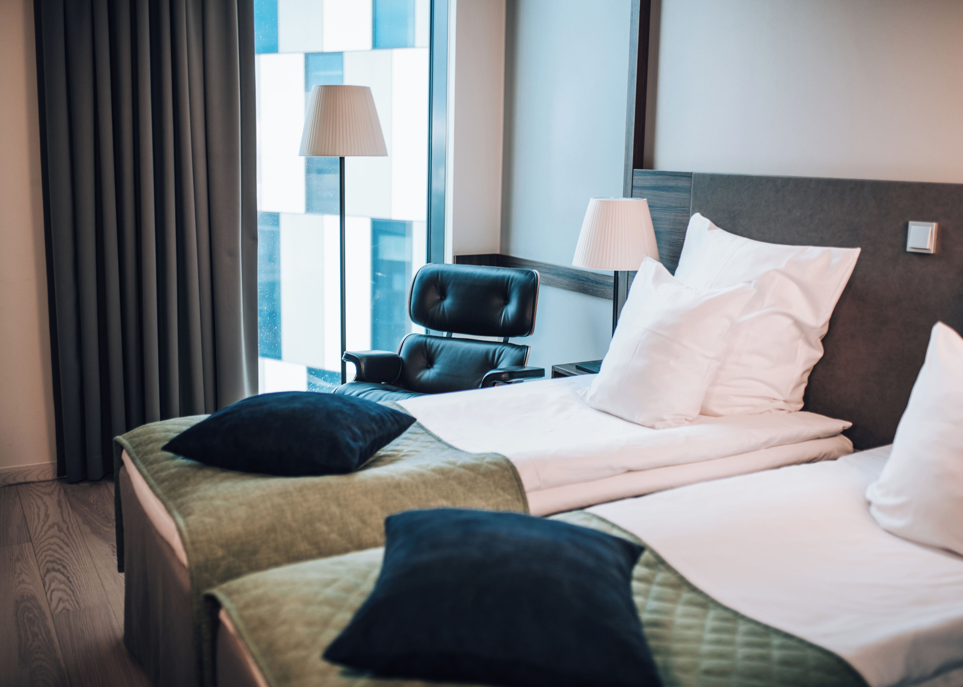A hotel room with a bed, pillows, a chair, a nightstand, a lamp, and a window with curtains.