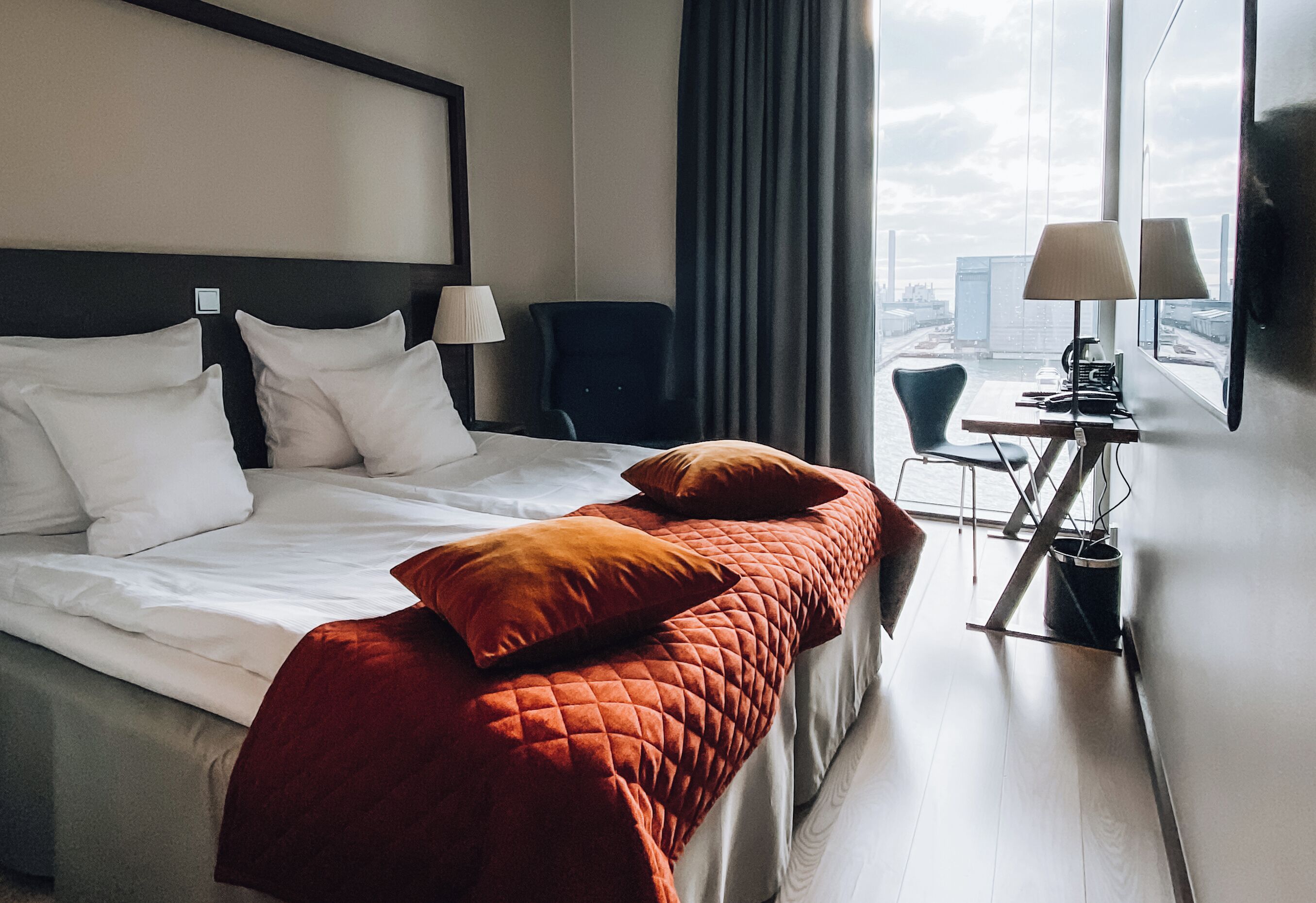 Una habitación de hotel con una cama, dos sillas, un escritorio con lámpara y vistas a la ciudad.