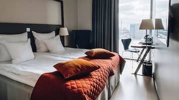Una habitación de hotel con una cama, dos sillas, un escritorio con lámpara y vistas a la ciudad.