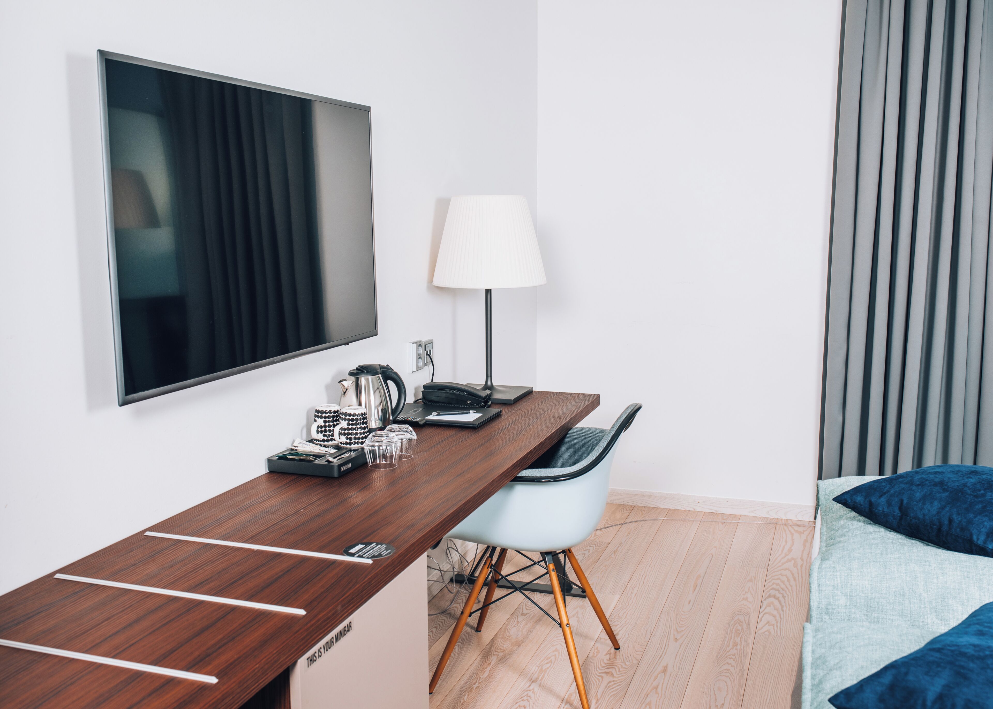 Une chambre d’hôtel moderne avec un bureau en bois, un téléviseur à écran plat, une lampe, une chaise et un lit avec une literie bleue.