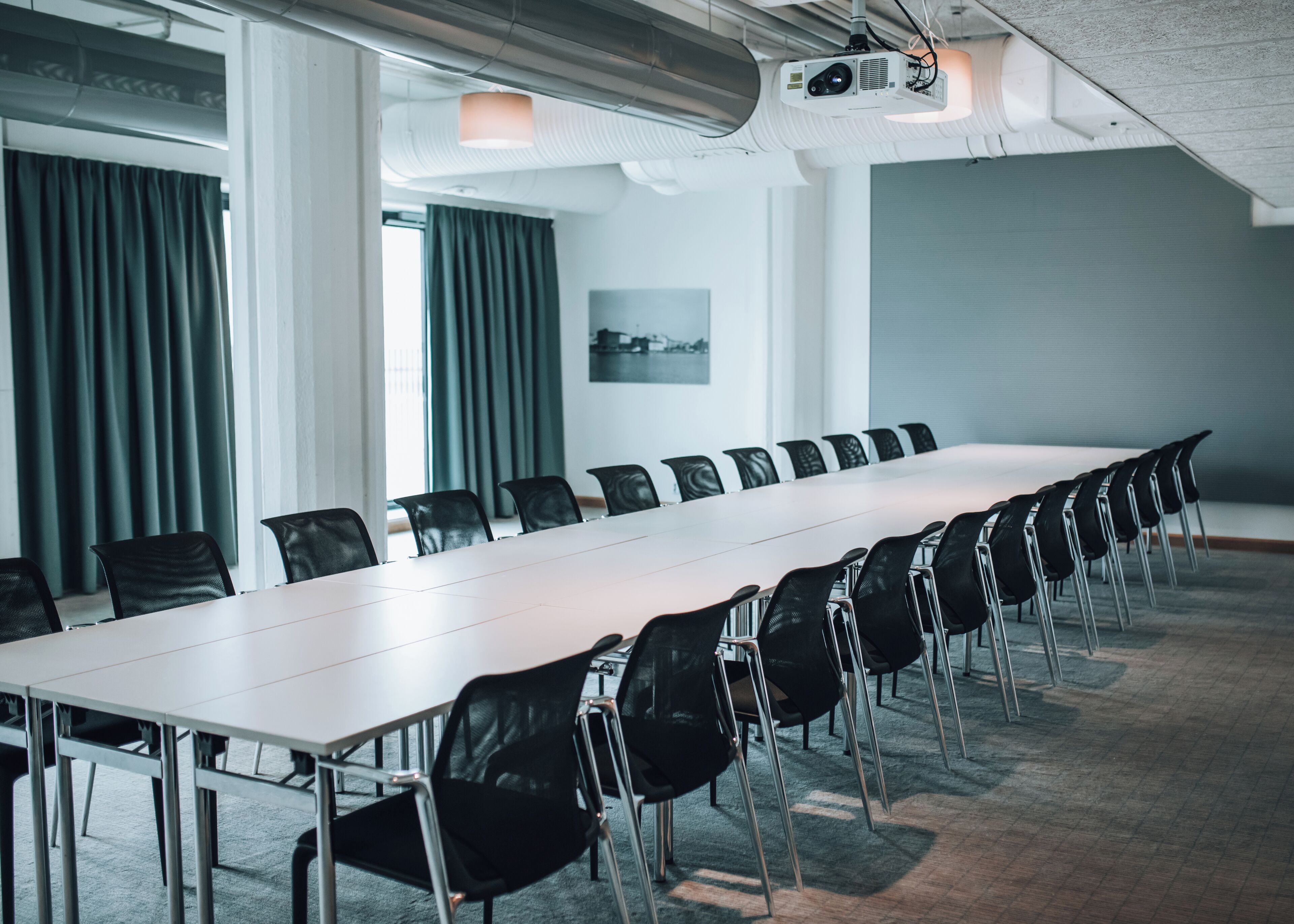 Une salle de conférence avec une longue table, des chaises noires, un projecteur et une grande fenêtre avec des rideaux.