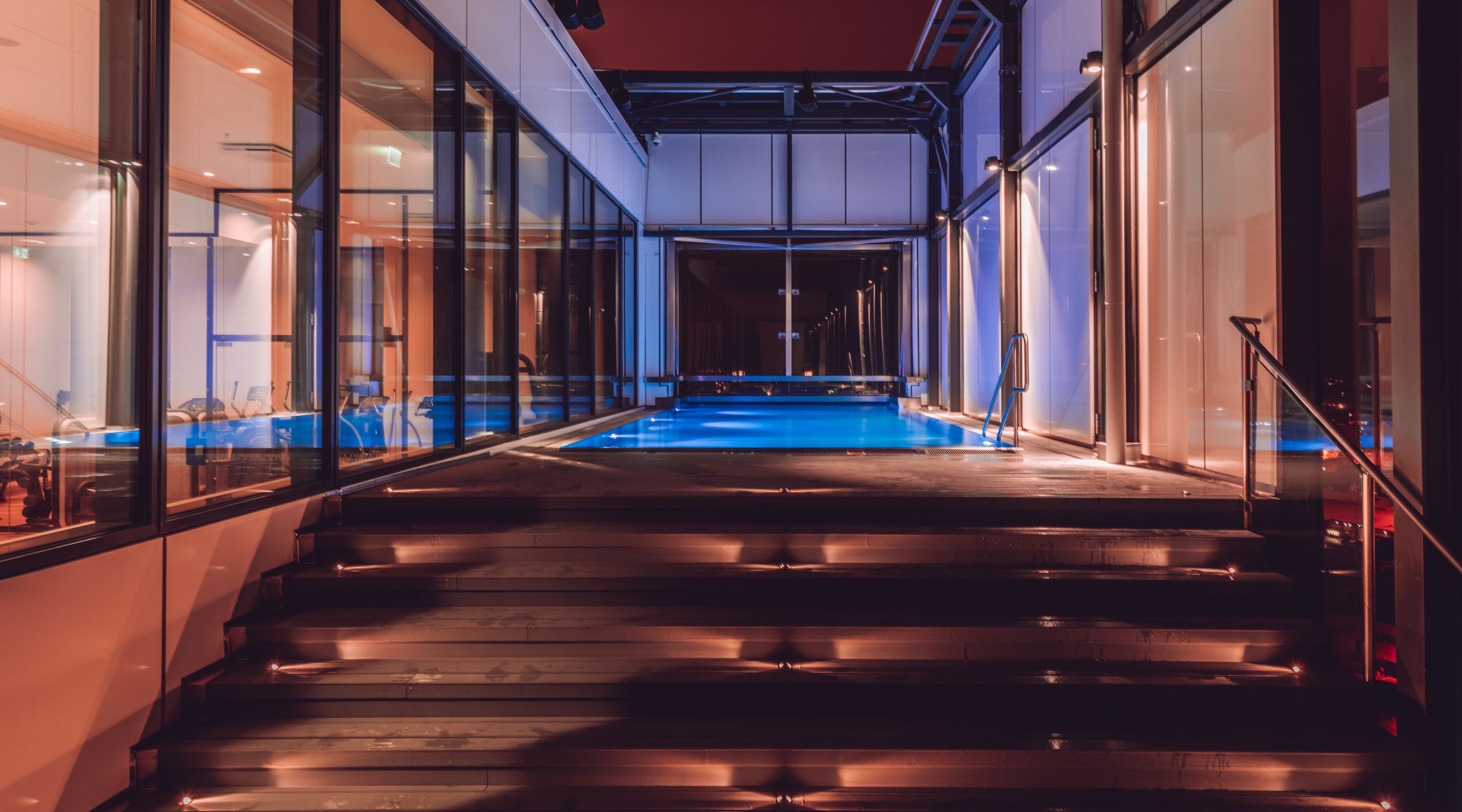 Zona de piscina interior moderna con paredes de vidrio y una escalera de madera que conduce a la piscina.