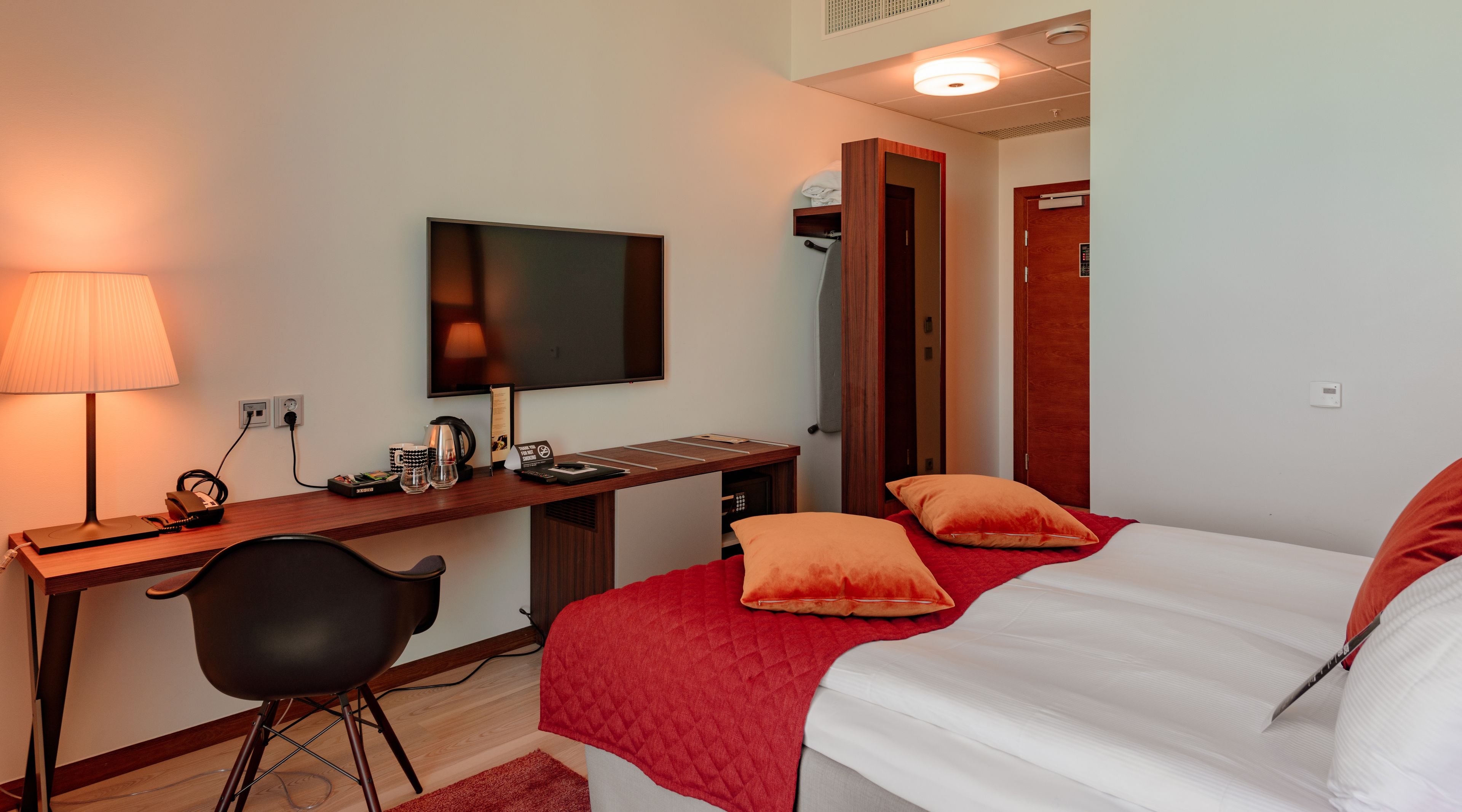 Una habitación de hotel con cama, escritorio con televisor, silla y colcha roja.