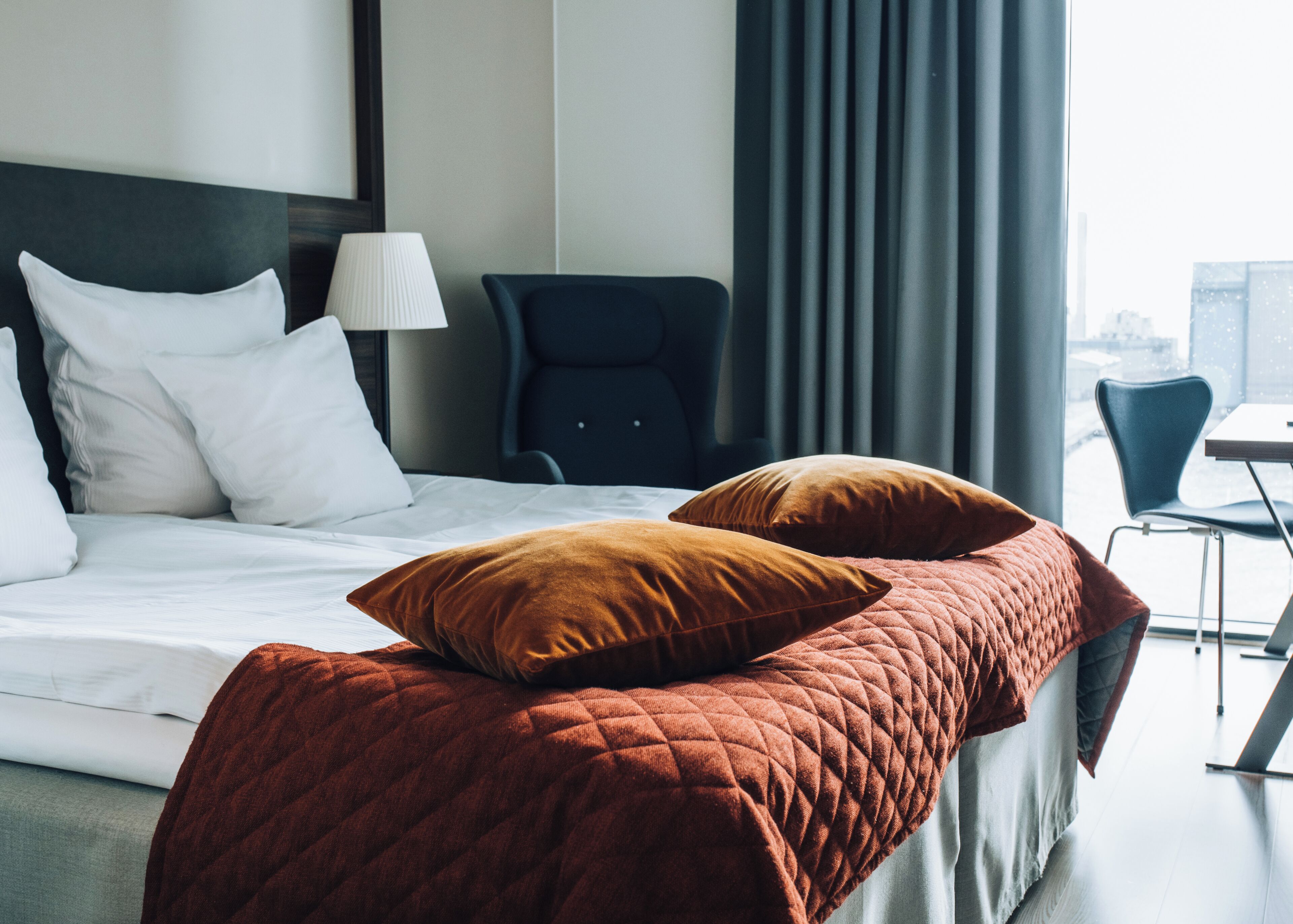 Une chambre d’hôtel avec un lit, des oreillers, une chaise, une lampe et une vue sur la ville par la fenêtre.