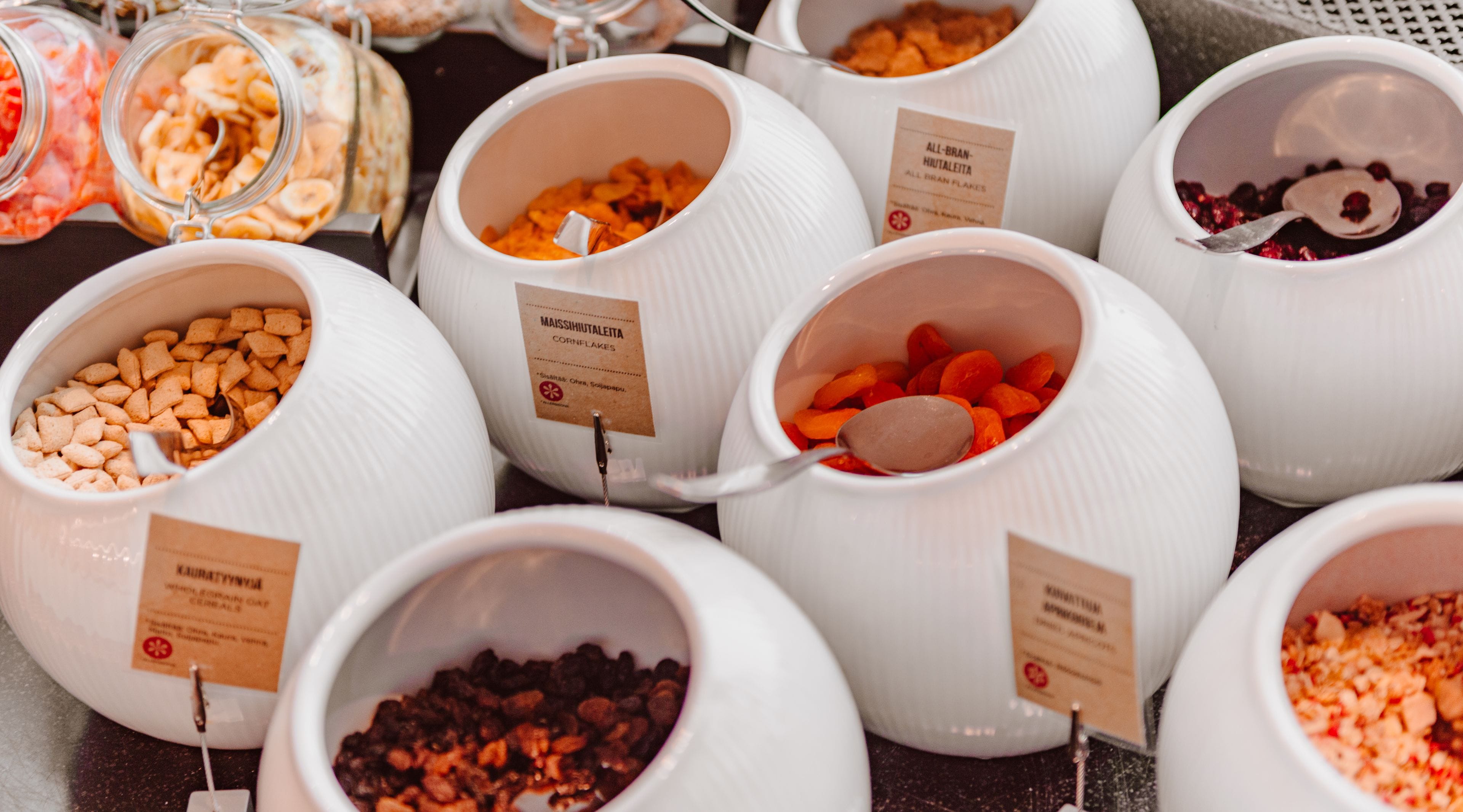 A variety of food items in white bowls with labels, including nuts, dried fruits, and cereal flakes.