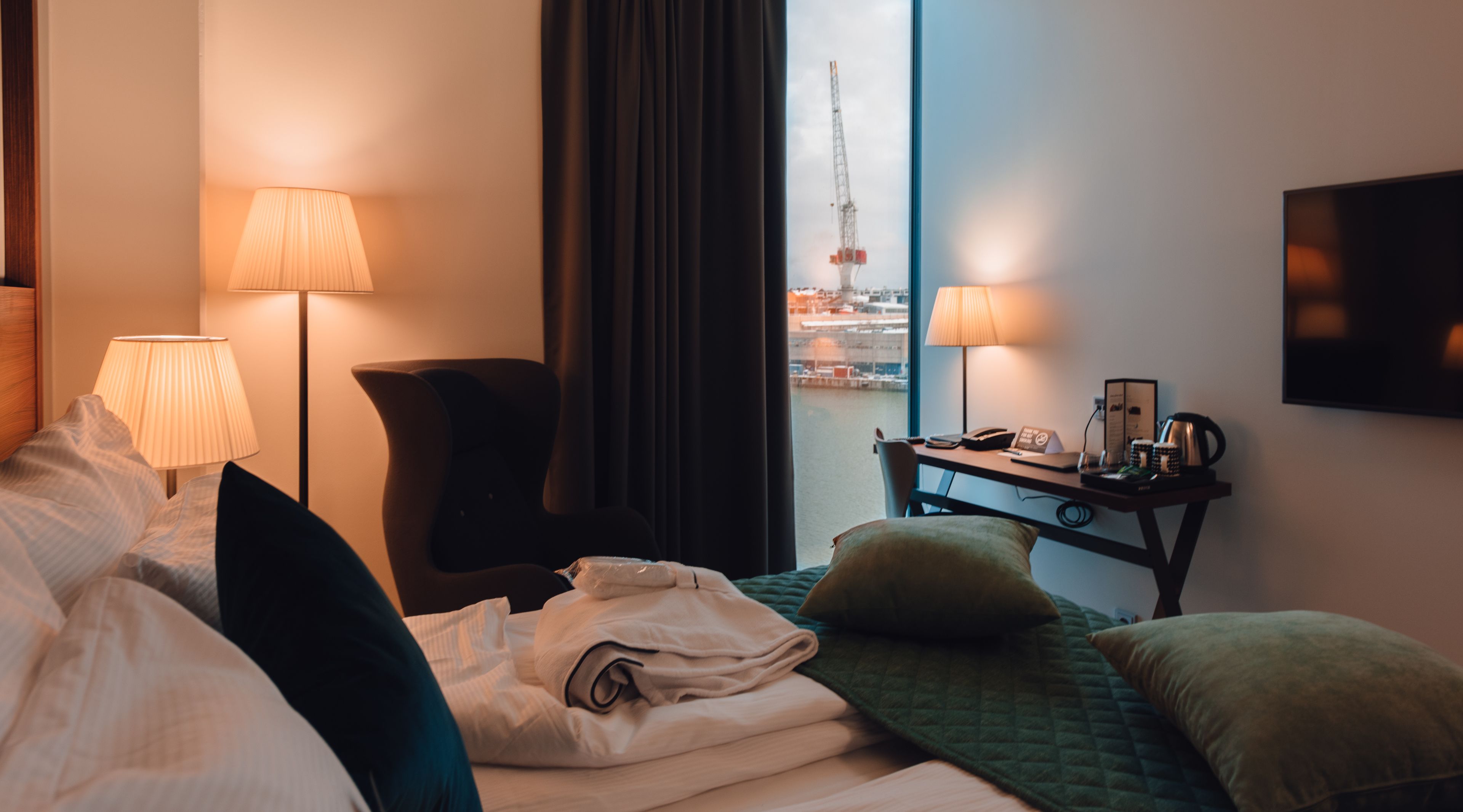 A hotel room with a bed, two bedside lamps, a desk with a lamp, a chair, and a view of a construction site through the window.