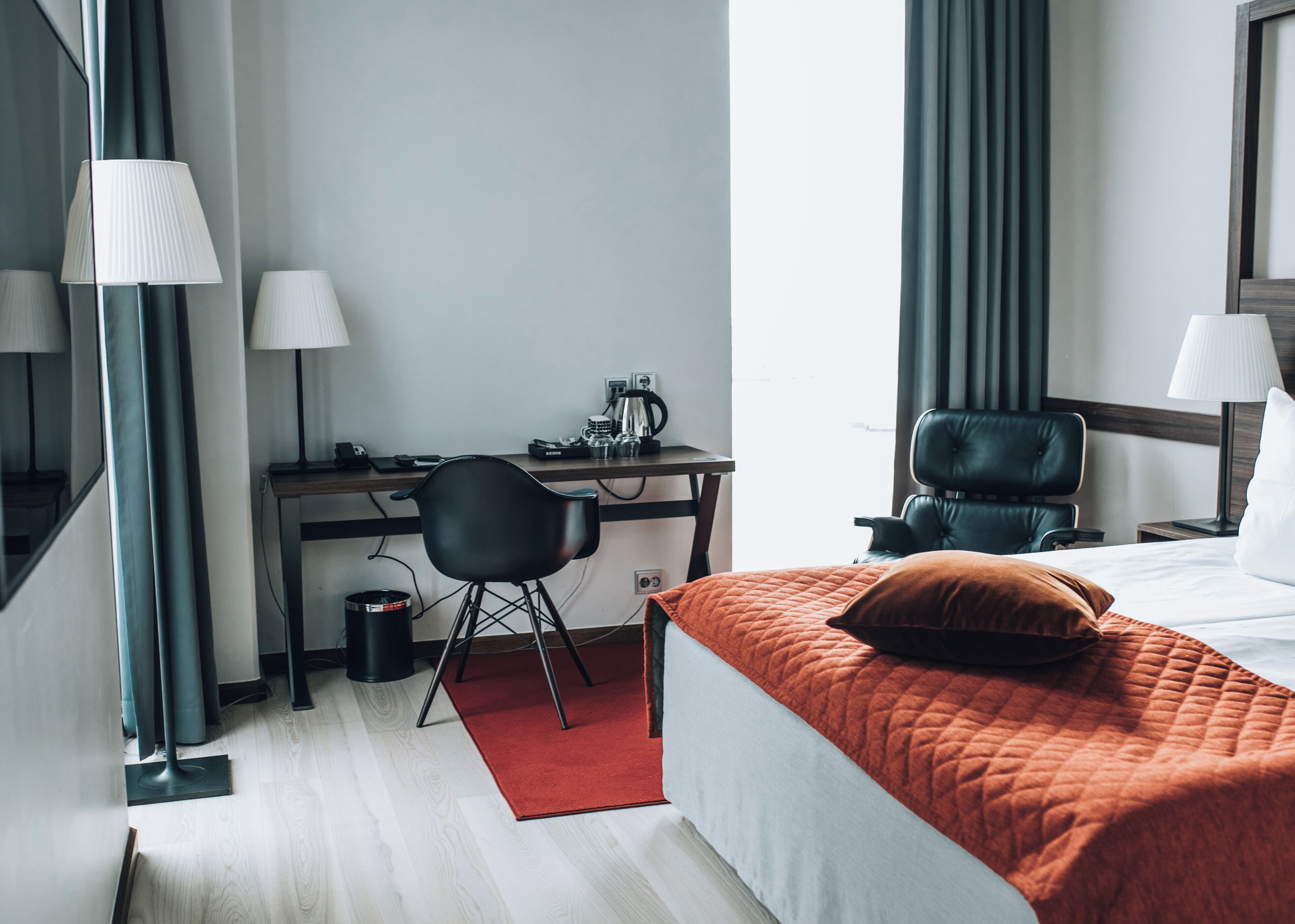 A hotel room with a bed, desk, chair, and a small trash bin.