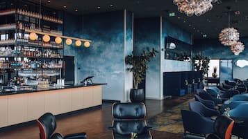 A modern bar area with shelves of bottles and glasses, a bar counter, and seating with a large potted plant.