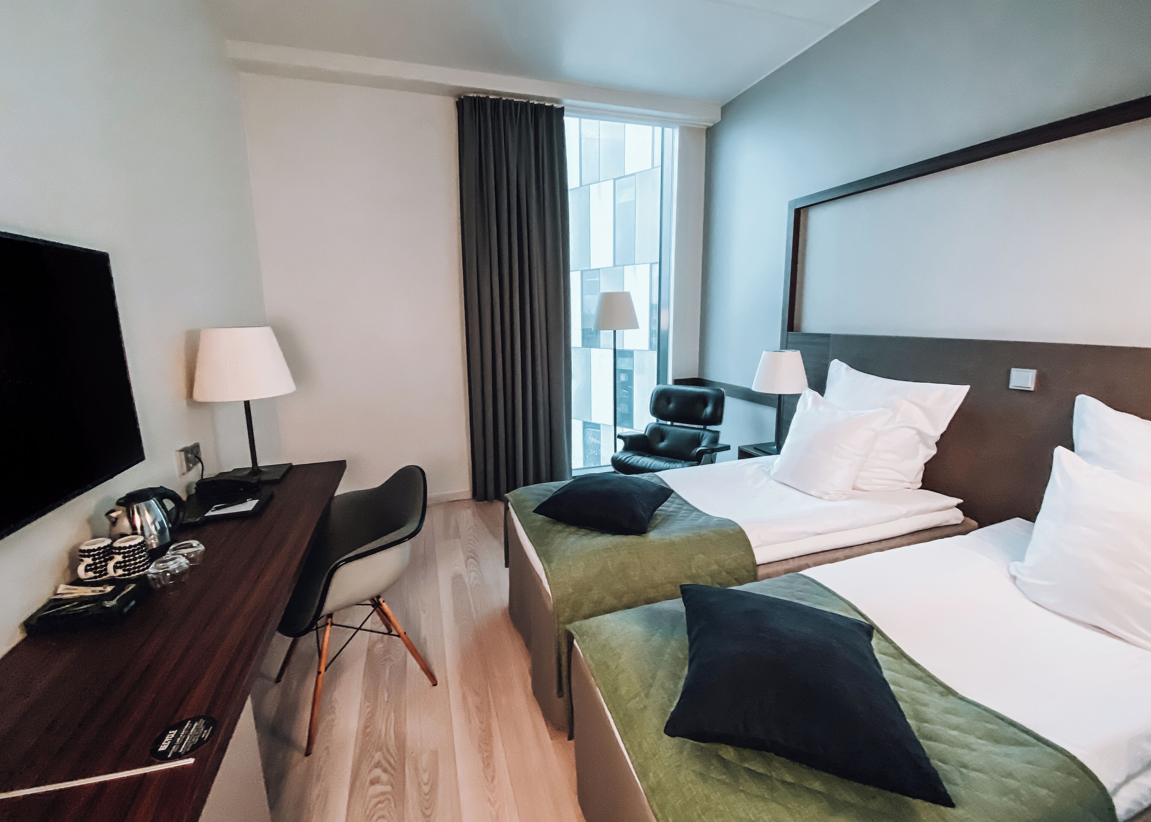 A hotel room with two beds, a desk, a chair, a TV, and a window with curtains.