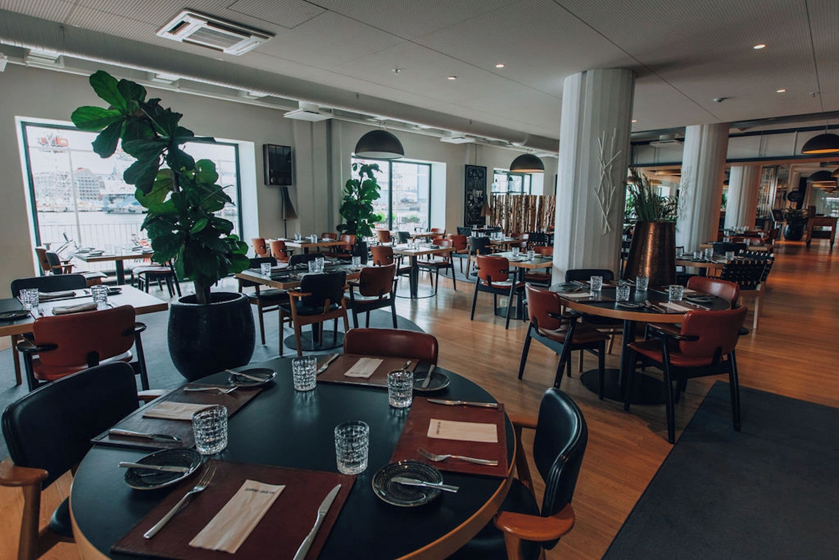 A spacious restaurant with multiple dining tables and chairs, large windows, and indoor plants.