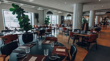 Un restaurante espacioso con varias mesas y sillas, ventanas amplias y plantas de interior.