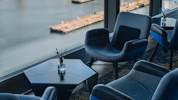 Un salón moderno con vistas a un río y al horizonte urbano, con asientos cómodos y una pequeña mesa con un adorno.