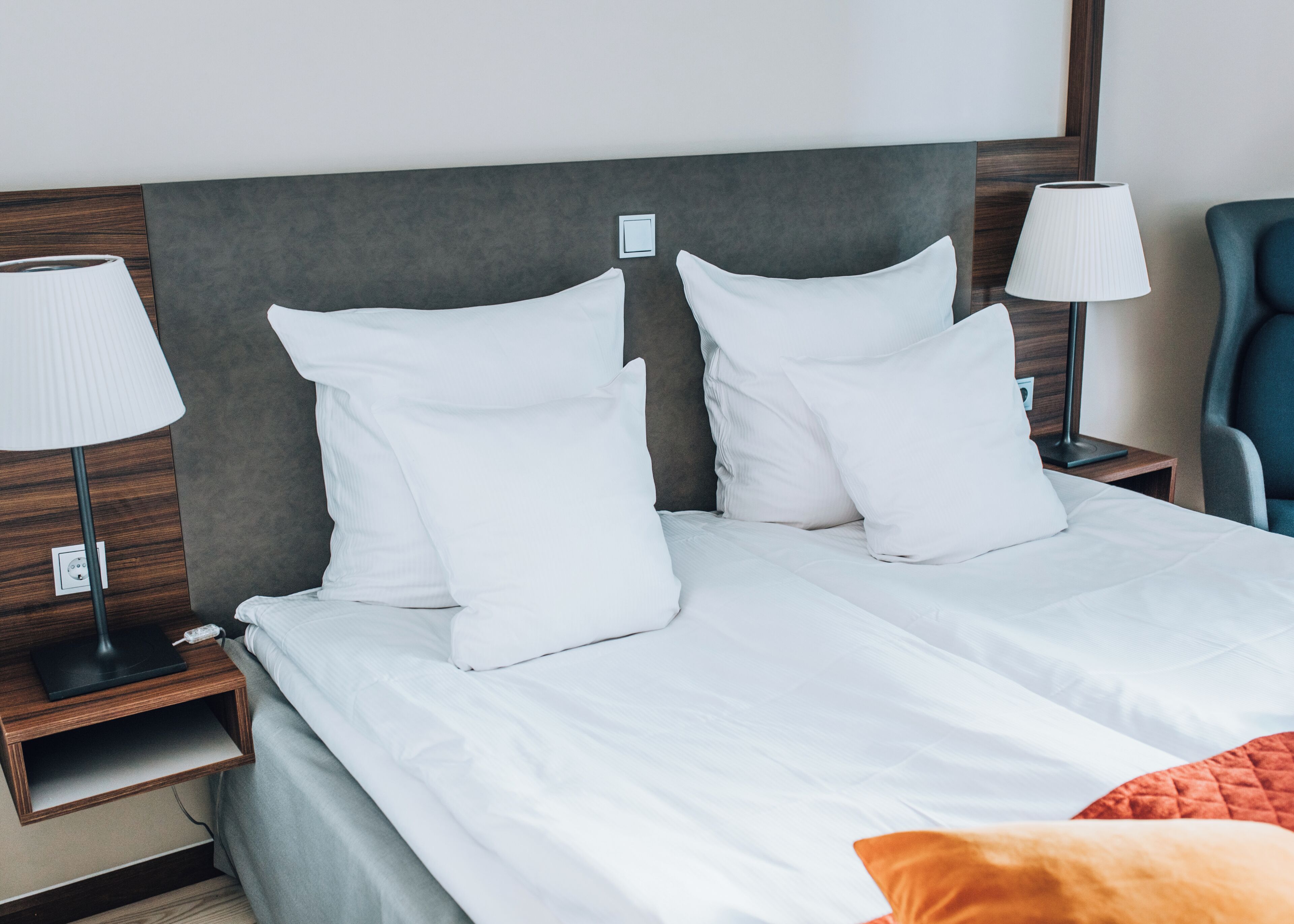 Una cama doble con ropa de cama blanca, dos mesitas de noche con lámparas, un cabecero de madera y una silla azul.