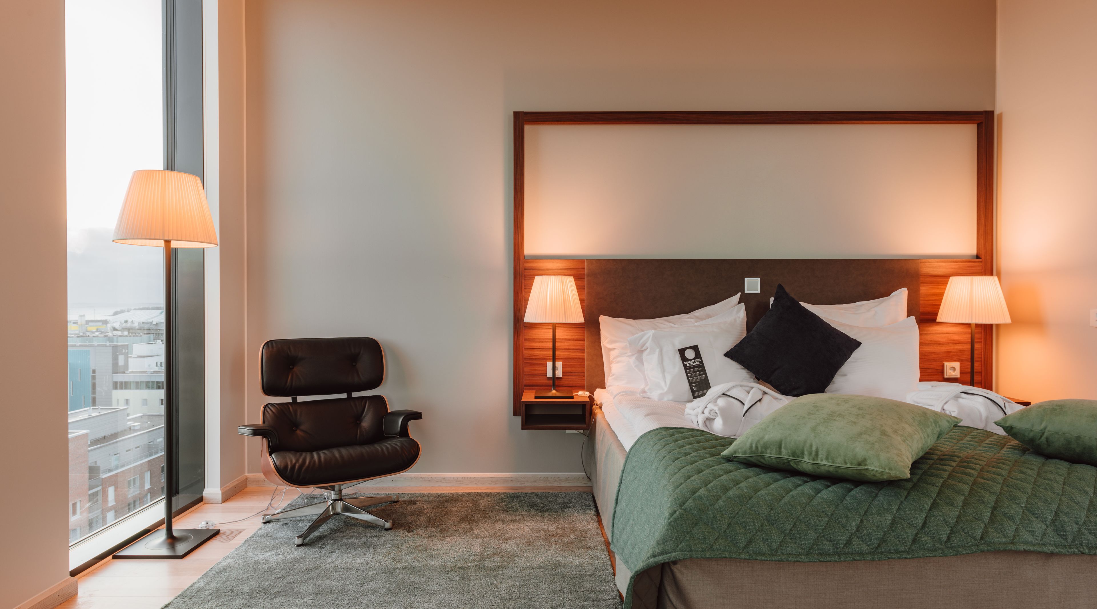A hotel room with a bed, two bedside lamps, a chair, and a view of the city through a large window.