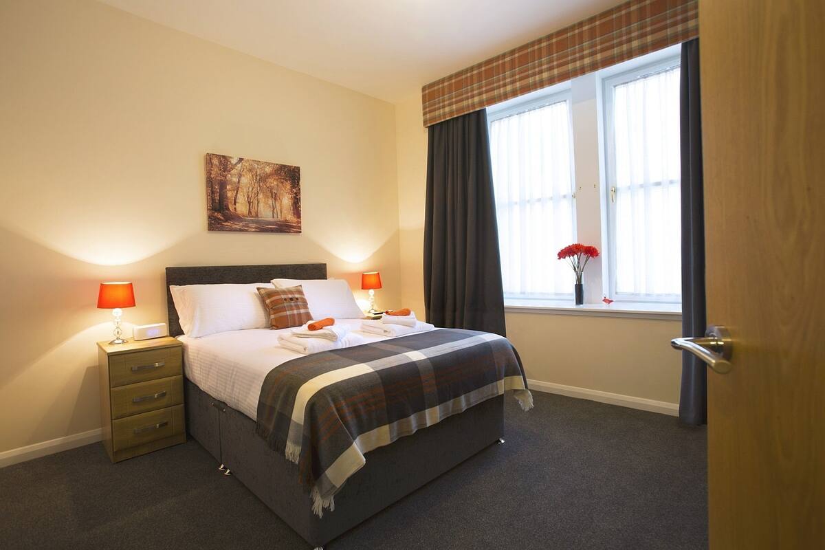 A neatly made bed with a plaid blanket, two bedside lamps, a nightstand, and a window with curtains.