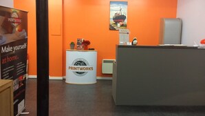 A reception area with an orange wall, a white reception desk, and a sign that reads "PRINTWORKS". - Printworks Apart-Hotel (Peterhead)