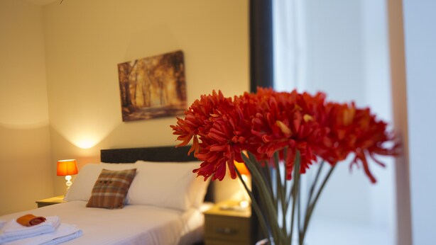 A hotel room with a bed, a nightstand with a lamp, a vase with flowers, and a painting on the wall. - Printworks Apart-Hotel (Peterhead)