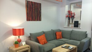 A modern living room with a grey sofa, two orange cushions, a wooden side table with a red lamp, and a small plant. - Printworks Apart-Hotel (Peterhead)