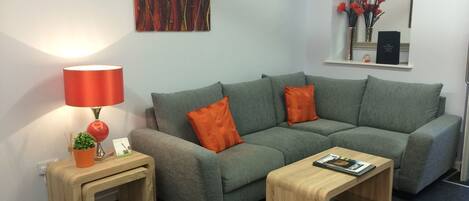 Una sala de estar moderna con un sofá gris, dos cojines naranja, una mesita de madera con una lámpara roja y una planta pequeña.